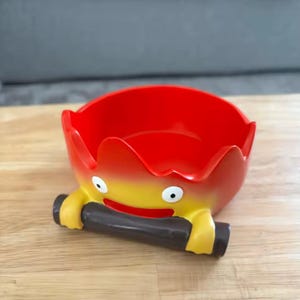 以下が含まれることがあります： 笑顔の顔と濃い茶色の丸太型のハンドルが付いた、赤と黄色の漫画スタイルのボウル。 ボウルの縁には炎のようなデザインがあります。