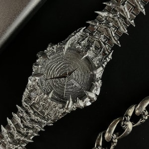 May include: A silver-toned wristwatch with a unique, textured design. The watch face has a circular pattern, and the band and bezel feature a spiky, irregular texture. The watch displays the day of the week. A silver chain is visible in the background.