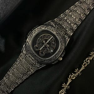 以下が含まれることがあります： 精巧な彫刻が施されたシルバーの腕時計。文字盤は暗く、装飾的なエンブレムとマッチするブレスレットが付いています。時計とブレスレットは暗い表面に表示されています。