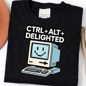 Puede incluir: Camiseta negra con un gráfico de ordenador retro y el texto "CTRL+ALT+DELIGHTED". El ordenador tiene una cara sonriente en la pantalla. La camiseta es de cuello redondo.