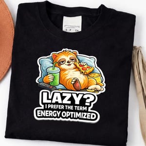 Könnte beinhalten: Schwarzes T-Shirt mit einem Cartoon-Faultier-Design. Das Faultier liegt auf einem Kissen, isst Pizza und trinkt aus einem grünen Becher. Der Text lautet: "LAZY? I PREFER THE TERM ENERGY OPTIMIZED."