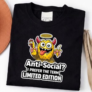 Könnte beinhalten: Schwarzes T-Shirt mit einem Cartoon-Monster, das ein Huhn und eine Banane hält. Das gelbe Monster hat Hörner und einen Heiligenschein. Der Text lautet: "Anti-Social? Ich bevorzuge den Begriff LIMITED EDITION."