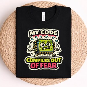May include: A black t-shirt featuring a graphic of a green, sweating computer processor character with wide eyes and glasses. The text reads "MY CODE" at the top and "COMPILES OUT OF FEAR" at the bottom in bold, colourful lettering with a white outline.