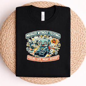 May include: A black t-shirt featuring a graphic of a light blue robot pressing a large enter key. The text reads "MERGED WITHOUT TESTING" and "FEELING CUTE, MIGHT REVERT" with computer error icons, a cursor, and a bug surrounding the central robot character.