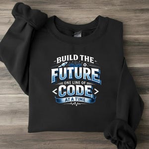 Op de afbeelding: Een zwart sweatshirt met ronde hals en een grafische print in blauwe en witte tinten. De tekst luidt BUILD THE FUTURE ONE LINE OF CODE AT A TIME, vergezeld van illustraties van een kleine raket, printplaatlijnen en haakjes.