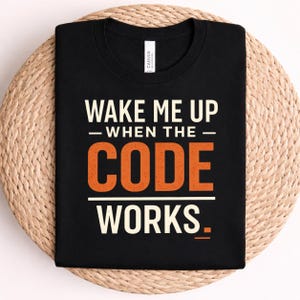 May include: A black crew neck t-shirt featuring the white and orange text WAKE ME UP WHEN THE CODE WORKS. The shirt has a white label at the inner neckline with the brand name Canvas visible. The design uses a mix of bold and distressed fonts.