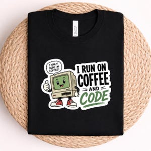 May include: A black t-shirt featuring a graphic of a smiling, anthropomorphic beige computer monitor wearing red trainers. The text reads I RUN ON COFFEE AND CODE, with a speech bubble saying I ONLY CODE IN COFFEE. The design uses black, green, and red.