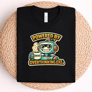 May include: A black t-shirt featuring a graphic of a smiling robot holding a mug with the word SYNTAX printed on it. Above the robot, the text reads POWERED BY, and below the robot, the text reads OVERTHINKING.EXE in orange and yellow lettering.