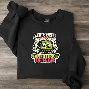 May include: A black crewneck sweatshirt featuring a graphic of a sweating, anxious green computer chip character with the text "MY CODE COMPILES OUT OF FEAR" printed in white and pink lettering above and below the central illustration.