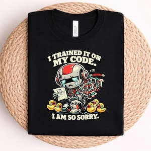May include: A black t-shirt featuring a graphic of a sad, crying robot with red and white accents, surrounded by yellow rubber ducks. The robot holds a paper that says I'M SORRY! Text above reads I TRAINED IT ON MY CODE. and below reads I AM SO SORRY.