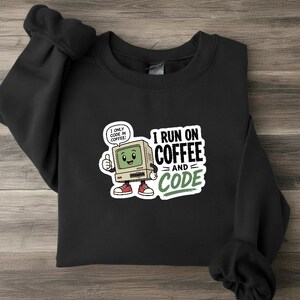 Pode incluir: Uma camisola preta de gola redonda com um gráfico de uma personagem de computador vintage com braços e pernas, fazendo um sinal de positivo. O texto diz I RUN ON COFFEE AND CODE, com um balão de fala dizendo I ONLY CODE IN COFFEE. O design usa detalhes em verde, branco e vermelho.
