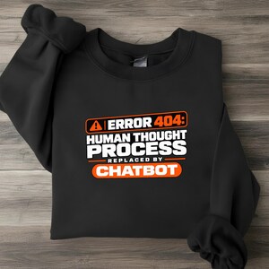 May include: A black crewneck sweatshirt featuring a graphic print with an orange warning icon and the text ERROR 404: HUMAN THOUGHT PROCESS REPLACED BY CHATBOT. The text is displayed in a mix of white and orange block lettering on a dark background.