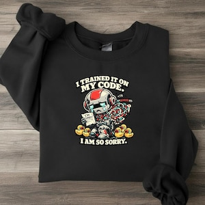 Könnte beinhalten: Ein schwarzes Sweatshirt mit Rundhalsausschnitt und einer Grafik eines weinenden Roboters mit freiliegenden bunten Kabeln, der einen Zettel mit der Aufschrift I'M SORRY! hält. Oben steht I TRAINED IT ON MY CODE. und unten I AM SO SORRY. mit gelben Enten.