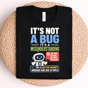 May include: A black t-shirt featuring a graphic with the text IT'S NOT A BUG IT'S A MISUNDERSTANDING. Below is a white robot with the letters AI on its chest, a red speech bubble stating I DID WHAT YOU SAID, NOT WHAT YOU MEANT, and a blue box with text.
