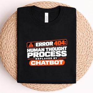 Könnte beinhalten: Ein schwarzes T-Shirt mit einer Grafik, die ein orangefarbenes Warnsymbol und den Text ERROR 404: HUMAN THOUGHT PROCESS REPLACED BY CHATBOT zeigt. Der Text ist in einer Mischung aus weißen und orangefarbenen Blockbuchstaben auf der Vorderseite.
