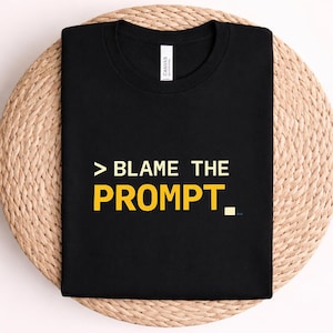 Könnte beinhalten: Ein schwarzes kurzärmeliges T-Shirt mit dem Text > BLAME THE PROMPT in weißen und gelben Buchstaben. Das T-Shirt hat einen Rundhalsausschnitt und ein weißes Markenetikett ist am inneren Kragenbereich auf dem schwarzen Stoff zu sehen.