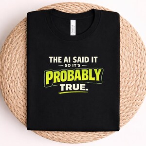 Op de afbeelding: Een zwart t-shirt met korte mouwen en de witte en limoengroene tekst THE AI SAID IT SO IT'S PROBABLY TRUE. Het shirt heeft een ronde hals en een wit merklabel zichtbaar bij de binnenkraag, geplaatst op een neutrale geweven ondergrond.