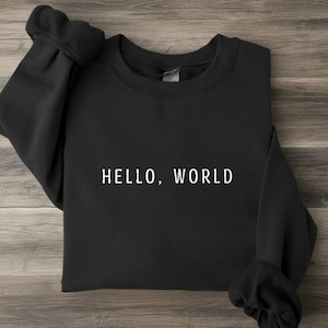 May include: A black long-sleeve crewneck sweatshirt featuring the text HELLO, WORLD printed in white capital letters across the center of the chest. The garment has a ribbed collar, cuffs, and hem, with a simple and clean design.