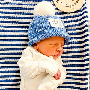 May include: A newborn baby wearing a blue knitted hat with a white pom-pom and a name tag that says "Rory" is sleeping on a blue and white striped blanket. A wooden sign with the name "Rory" and the word "girl" is in the background.
