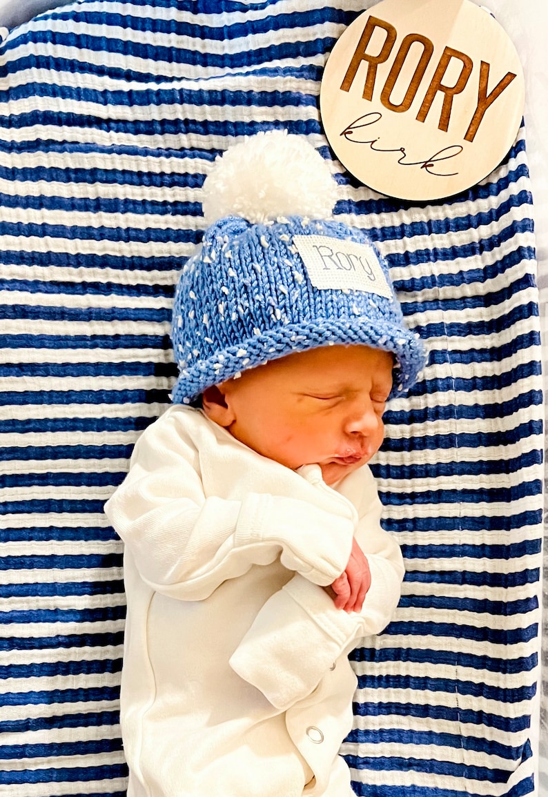May include: A newborn baby wearing a blue knitted hat with a white pom-pom and a name tag that says "Rory" is sleeping on a blue and white striped blanket. A wooden sign with the name "Rory" and the word "girl" is in the background.