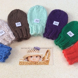 May include: Seven pairs of knitted baby mittens in various colours, including blue, beige, brown, green, purple, red, and light blue. Each mitten has a white tag with a number on it, ranging from 31 to 37.