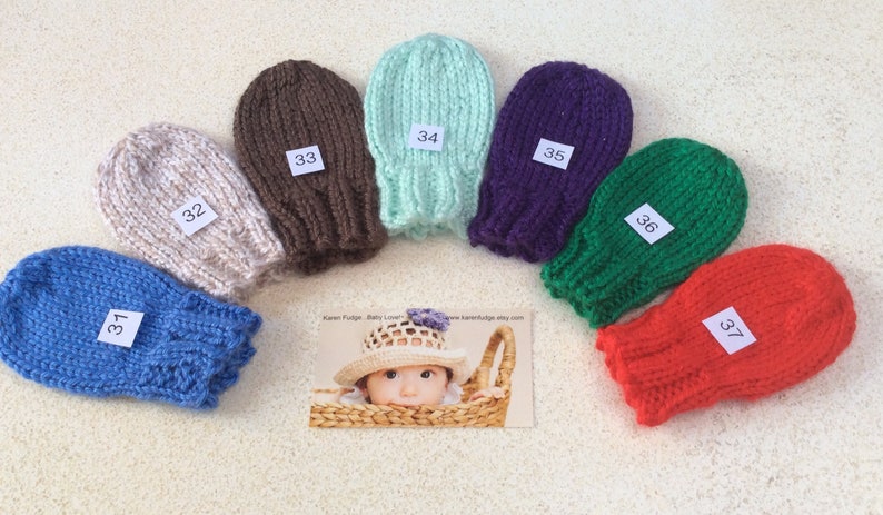May include: Seven pairs of knitted baby mittens in various colours, including blue, beige, brown, green, purple, red, and light blue. Each mitten has a white tag with a number on it, ranging from 31 to 37.
