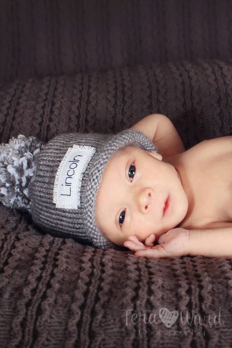 May include: A newborn baby wearing a grey knitted hat with a white pom-pom and a name tag that reads "Lincoln". The baby is lying on a brown knitted blanket.