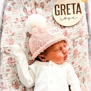 May include: A newborn baby girl wearing a pink knitted hat with a white pom-pom and a white onesie. The hat has the name "Greta" embroidered on it. A wooden sign with the name "Greta Rose" is in the background.