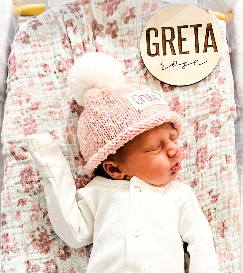 May include: A newborn baby girl wearing a pink knitted hat with a white pom-pom and a white onesie. The hat has the name "Greta" embroidered on it. A wooden sign with the name "Greta Rose" is in the background.