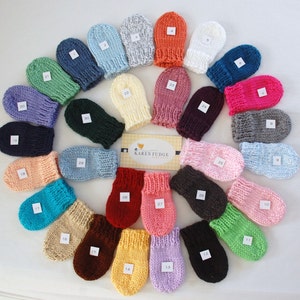 May include: A collection of 30 knitted baby mittens in various colours, including pink, blue, green, yellow, and red. The mittens are arranged in a circle, with the brand name "KAREN FUDGE" in the centre.