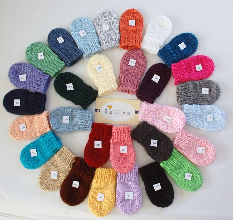 May include: A collection of 30 knitted baby mittens in various colours, including pink, blue, green, yellow, and red. The mittens are arranged in a circle, with the brand name "KAREN FUDGE" in the centre.