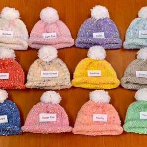 May include: Twelve knitted baby hats with pom-poms in various colours and patterns, including rainbow, light pink, violet, powder blue, salmon, sweet cream, sunshine, silver lining, blue lapis, sweet pink, fresh mint, and peachy.