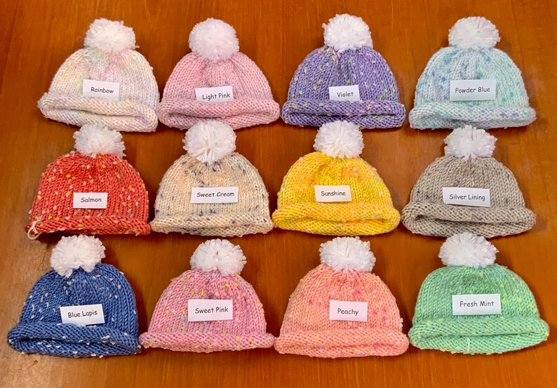 May include: Twelve knitted baby hats with pom-poms in various colours and patterns, including rainbow, light pink, violet, powder blue, salmon, sweet cream, sunshine, silver lining, blue lapis, sweet pink, fresh mint, and peachy.