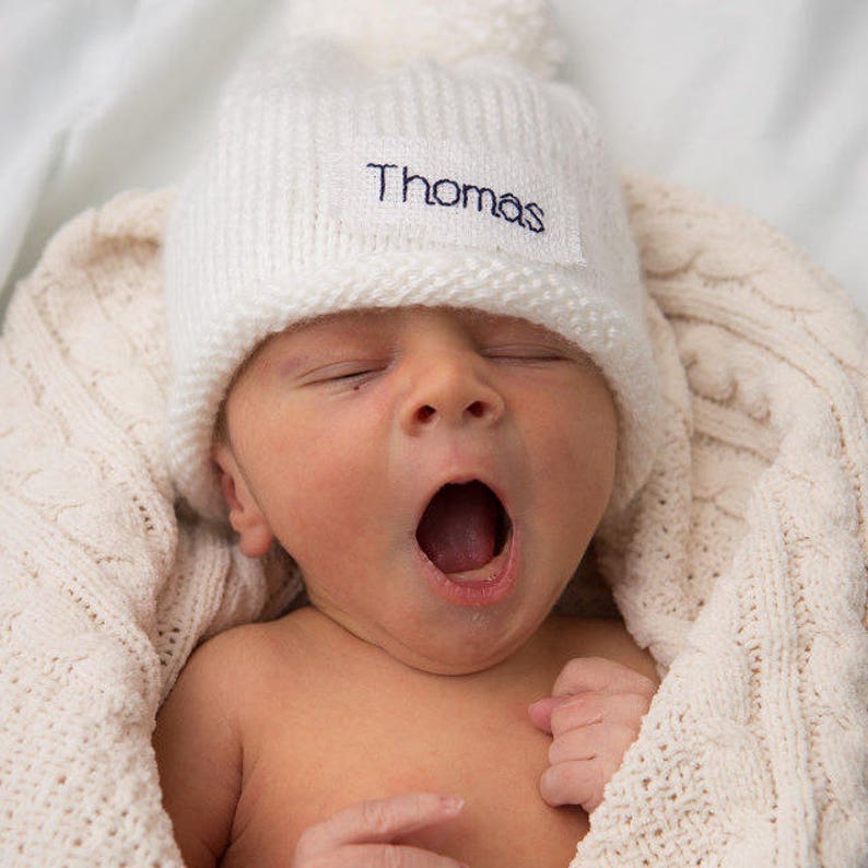 May include: A newborn baby wearing a white knitted beanie with the name "Thomas" embroidered on it. The baby is wrapped in a white knitted blanket and is yawning.