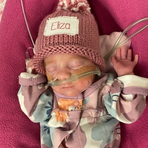 May include: A newborn baby girl wearing a pink knitted hat with a white pom-pom and a name tag that reads "Eliza". The baby is wearing a pink and blue floral onesie and has a clear nasal cannula.