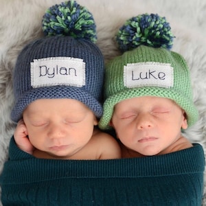May include: Two newborn babies wearing knitted hats with pom-poms. The hats are personalised with the names "Dylan" and "Luke". The babies are sleeping and wrapped in green blankets.