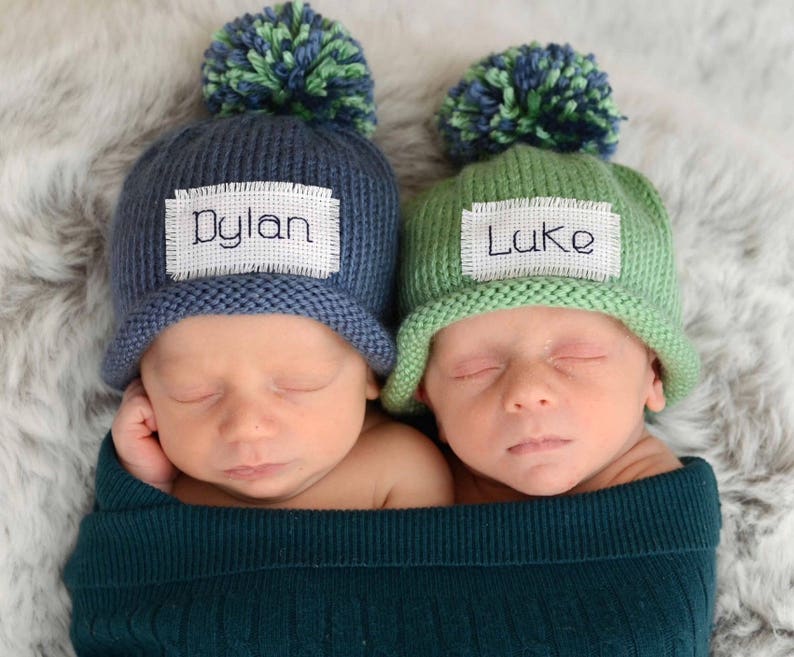 May include: Two newborn babies wearing knitted hats with pom-poms. The hats are personalised with the names "Dylan" and "Luke". The babies are sleeping and wrapped in green blankets.