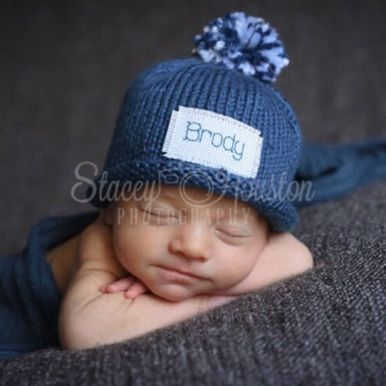 May include: A blue knitted baby hat with a white pom pom and a white label that says "Brody" in blue embroidery. The hat is on a baby's head.