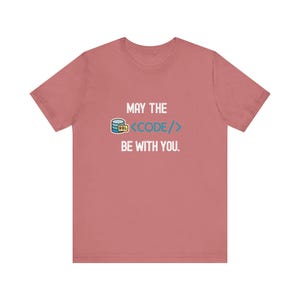 Può includere: T-shirt rosa antico con la scritta "MAY THE CODE BE WITH YOU". Il design include un'icona del database e parentesi di codice. Ideale per programmatori e appassionati di coding.