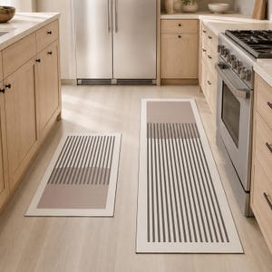May include: Two rectangular kitchen floor mats featuring a geometric pattern of vertical dark grey stripes over a light beige background. The mats have a clean border and are positioned on a light wood floor in a kitchen with light wood cabinets.