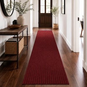 May include: A long, rectangular runner rug featuring a deep red base color with thin, vertical black stripes running the entire length of the fabric. The rug is positioned centrally on a dark hardwood floor in a hallway setting.