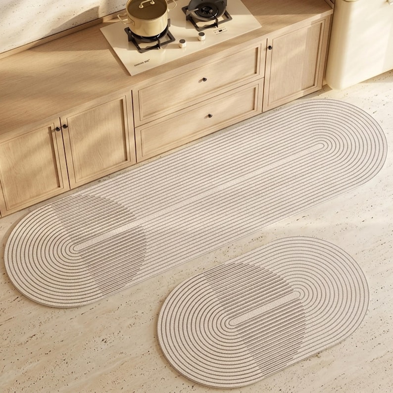 May include: A set of two oval-shaped floor mats featuring a light beige and white linear pattern of concentric rounded lines. The mats are positioned on a light-coloured floor in front of wooden kitchen cabinetry and a stovetop with a pot.