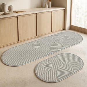 May include: A set of two oval-shaped floor mats featuring a light grey and white linear pattern of concentric semicircles and straight lines. The mats have a smooth, low-profile surface with a consistent geometric design across both pieces.