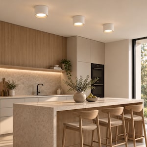 Könnte beinhalten: Drei weiße, zylindrische Deckenleuchten mit warmer Beleuchtung, die in einer Reihe installiert sind. Diese modernen Leuchten zeichnen sich durch ein schlichtes, kreisförmiges Design aus, das den Küchenbereich direkt nach unten beleuchtet.