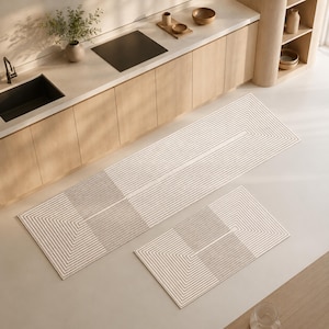 May include: A set of two rectangular floor mats featuring a geometric pattern of concentric lines in shades of beige and dark grey. The mats are arranged on a light-coloured floor in front of kitchen cabinetry, displaying a repeating linear design.