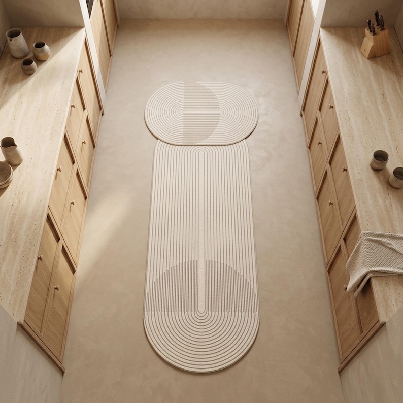 May include: Two elongated oval rugs with a cream and beige linear pattern featuring concentric semi-circles and straight lines. The rugs are positioned on a light-coloured floor between two sets of wooden cabinets with light-coloured countertops.