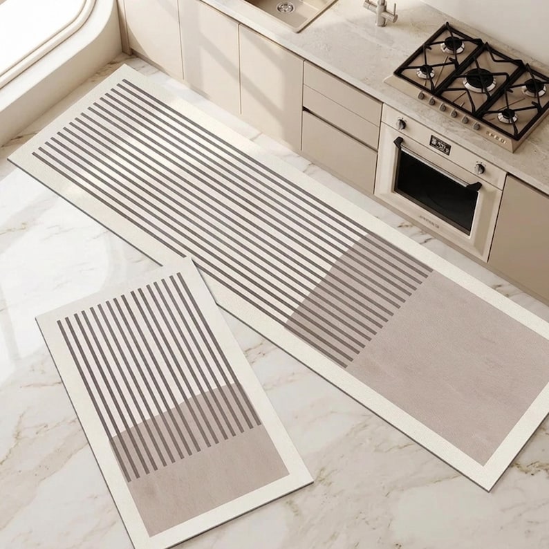 May include: A set of two rectangular kitchen floor mats featuring a cream-coloured border and a pattern of thin, dark brown vertical stripes that transition into a solid light brown block at one end of each mat, creating a clean, linear design.