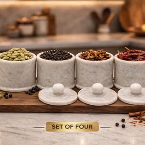 May include: A set of four white marble spice jars filled with green cardamom, black peppercorns, dried herbs, and red chili peppers, displayed on a wooden tray. The jars have matching lids with white spherical knobs. The text "SET OF FOUR" is visible.