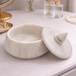Op de afbeelding: Een witte marmeren sieradendoos met een gedeeltelijk open deksel. Het deksel heeft een conische handgreep. De doos staat op een licht oppervlak, mogelijk een kaptafel. Het ontwerp is minimalistisch en elegant.