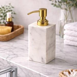 May include: A white marble soap dispenser with a gold pump. The rectangular dispenser is on a marble countertop. The background features a wooden tray, a glass vase, and a stack of white towels.
